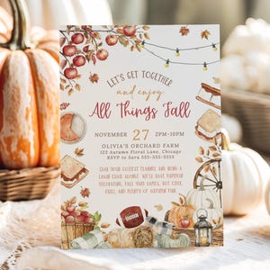 Rustic All Things Fall party invitation featuring watercolor autumn illustrations including apples on branches, pumpkins, falling leaves, lantern, hay bale and cozy fall foods surrounding warm seasonal lettering. Soft earth tone typography highlights the editable event details in the center. The card stands upright on a wooden surface with pumpkins and woven textures in the background creating a warm autumn harvest atmosphere.