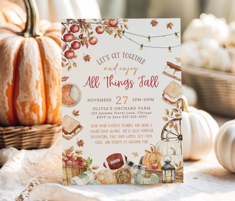 Rustic All Things Fall party invitation featuring watercolor autumn illustrations including apples on branches, pumpkins, falling leaves, lantern, hay bale and cozy fall foods surrounding warm seasonal lettering. Soft earth tone typography highlights the editable event details in the center. The card stands upright on a wooden surface with pumpkins and woven textures in the background creating a warm autumn harvest atmosphere.