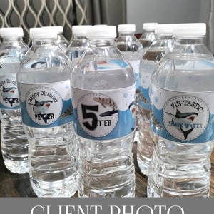 May include: A group of clear plastic water bottles with personalised labels featuring a shark and the text "Happy Birthday Peter" and "Fin-Tastic Shark Water".