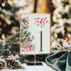 May include: A rectangular table number sign in a gold frame featuring a watercolor illustration of a Christmas tree with pink gifts and the red text Table 1 in a large green font. The design includes small hanging ornaments.