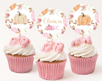 Può includere: Tre cupcake rosa e bianchi con glassa bianca e dettagli rosa. Ogni cupcake ha un topper rotondo di carta bianca con un design ad acquerello di zucche, foglie e fiori. Il topper sul cupcake centrale dice "Olivia is One" in lettere arancioni e rosa.