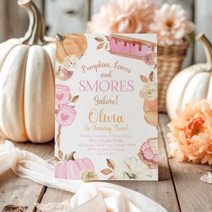 Printable Pumpkin and Smores Birthday Invitation styled on weathered wooden planks beside an oversized white pumpkin, peach garden blooms, woven baskets, and flowing ivory ribbon. The rustic farmhouse setting enhances the watercolor pumpkins, pink marshmallows, toasted smores, and soft floral artwork while showcasing this editable Canva invitation for a cozy autumn birthday party.