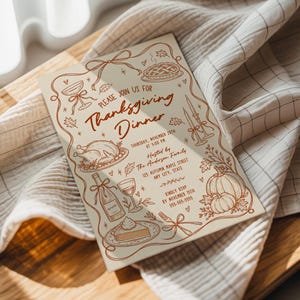 Hand drawn Thanksgiving invitation resting on soft neutral linens beside a warm wood surface. Brown illustrated details include a Thanksgiving turkey, pie, pumpkins, wine bottle, candles, autumn leaves and whimsical bows. Natural sunlight falls across the invitation and fabric. The cozy setting complements the casual Thanksgiving dinner theme.