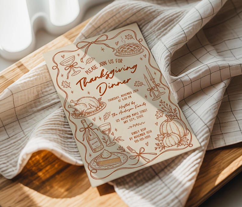 Hand drawn Thanksgiving invitation resting on soft neutral linens beside a warm wood surface. Brown illustrated details include a Thanksgiving turkey, pie, pumpkins, wine bottle, candles, autumn leaves and whimsical bows. Natural sunlight falls across the invitation and fabric. The cozy setting complements the casual Thanksgiving dinner theme.