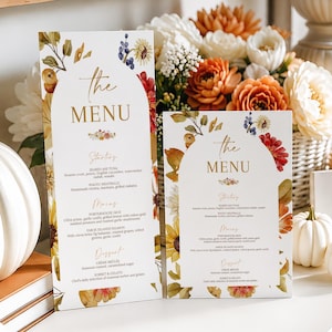 Könnte beinhalten: Zwei weiße, rechteckige Menükarten mit bogenförmigen oberen Rändern, verziert mit goldenem Text und floralen Aquarellillustrationen. Der Text enthält Überschriften für Starters, Mains und Dessert sowie Gerichte wie Seared Ahi Tuna und Wagyu Meatballs.