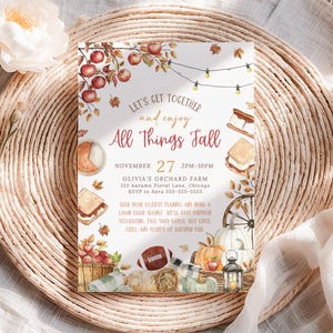 Autumn harvest party invitation with watercolor apples, s’mores, pumpkins and string lights framing the All Things Fall headline in warm rustic colors. Editable party details appear in the center with soft seasonal typography. The invitation sits on a woven placemat beside neutral florals and soft textured fabric adding an elegant farmhouse fall aesthetic.