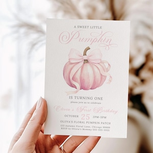 Fall-themed pink pumpkin birthday invitation for a girl's first birthday featuring a watercolor pink pumpkin with a pale pink coquette bow. The design is clean and feminine with a vintage flair. The card is held in hand and printed on textured cardstock, captured in soft natural light with a neutral blurred background, creating a warm and gentle tone ideal for showcasing the physical print quality.