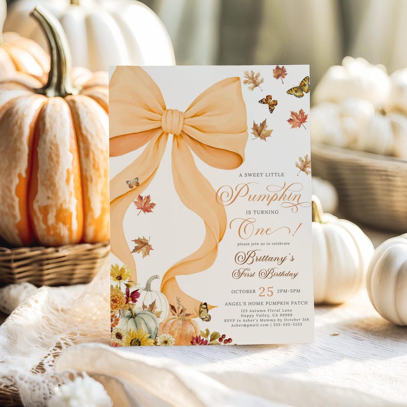 Pastel pumpkin and coquette bow first birthday invitation featuring watercolor pumpkins, autumn leaves, and butterflies, styled upright against a wicker basket backdrop surrounded by cream and orange pumpkins. Ideal for a girl’s rustic fall birthday celebration, pastel autumn party, or boho pumpkin patch birthday with customizable editable party details.