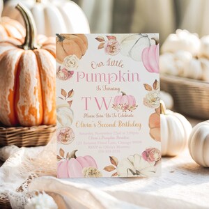 The Printable Occasion second birthday invitation with a pumpkin theme, perfect for fall or Thanksgiving celebrations. Design includes pink, white, and orange watercolor pumpkins with delicate florals. Text reads “Our Little Pumpkin is Turning Two.” Mockup shown standing upright among real pumpkins in a rustic outdoor basket display, highlighting an autumn harvest theme.