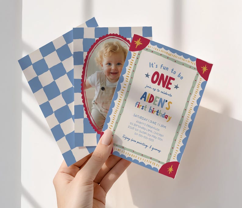 Holding a Fun To Be One first birthday invitation, a hand presents the colorful card alongside the matching checkerboard photo back. Bright vintage inspired typography and whimsical hand drawn elements frame the party details. The reverse side features a personalized child photo within a decorative oval frame. Natural window light casts soft shadows across the scene for an airy modern look.