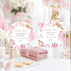 May include: Two spa-themed ornaments with pink watercolor illustrations. One is round, the other square. Both feature a robe, eye mask, and text: "Thank You For Making My Party Spa-tacular." A pink towel and makeup are in the foreground.