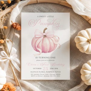Printable fall birthday invitation featuring a pastel pink watercolor pumpkin with an oversized soft ribbon bow, styled for a girl's first birthday. The invite lies on a rattan basket liner with cream fabric, white mini pumpkins, and burnt orange dried florals, creating a cozy and whimsical autumn vibe. This feminine, vintage-inspired scene highlights the soft seasonal color palette of the design.