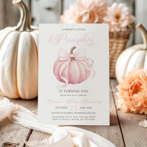 Editable girl’s pink pumpkin first birthday invitation placed among soft peachy florals and white pumpkins. The artwork features a large pink pumpkin with a flowing pastel bow and elegant fonts. Styled in a bright and airy flat lay with white drapery and wooden texture peeking through, this photo has a light coquette cottagecore vibe that blends baby girl charm with autumn elegance.