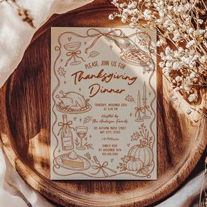 Thanksgiving dinner party invitation displayed on a rich wood serving board surrounded by soft cream fabric and dried flowers. The cream invitation features warm brown hand drawn turkey, pie, pumpkin, wine, candle and autumn leaf illustrations. Sunlight creates soft shadows across the card. The styling gives the Friendsgiving or Thanksgiving invitation a warm seasonal presentation.