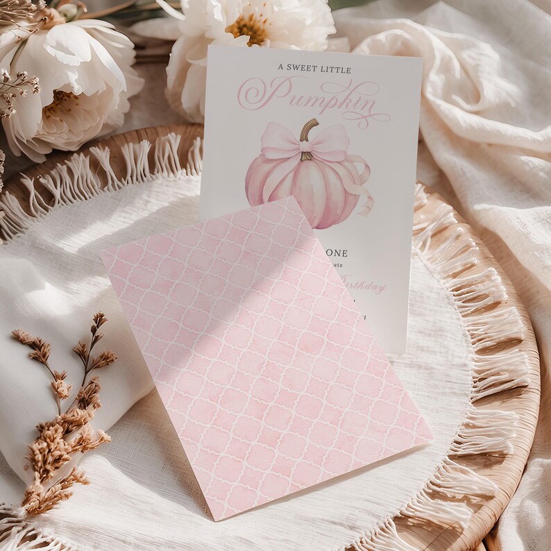 Back and front display of pink pumpkin 1st birthday invitation set for a girl’s fall party. The front features a watercolor pink pumpkin and bow design with elegant fonts, while the back shows a patterned soft pink quatrefoil print. Styled on a boho fringe woven tray with white florals and neutral linens, this mockup emphasizes the soft feminine tone and two-sided design versatility.