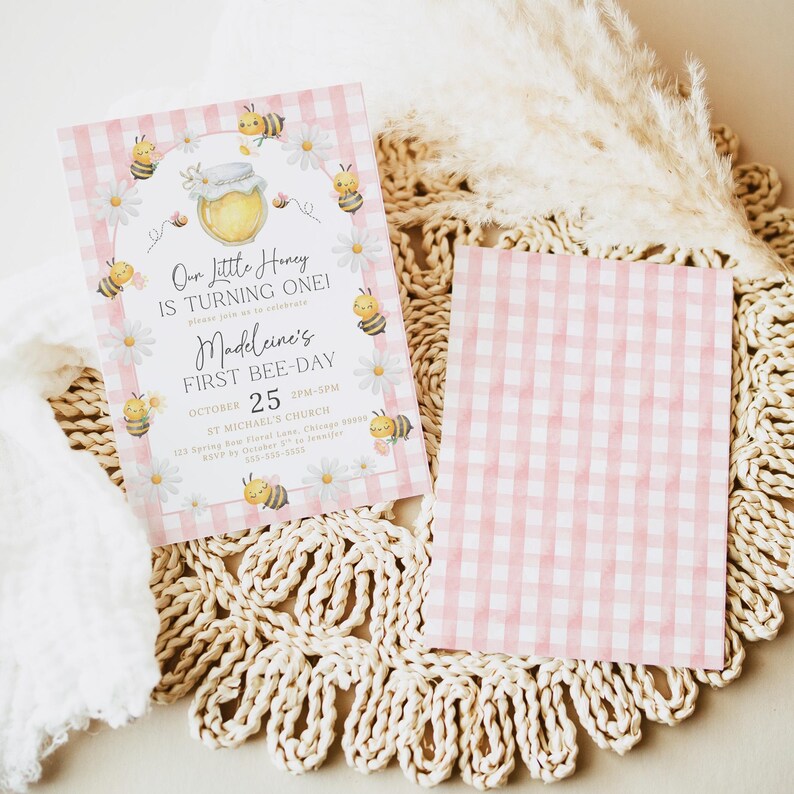 The Printable Occasions offers custom wedding birthday and baby shower templates for every celebration. Editable pink bee birthday invitation featuring watercolor bumblebees with pink wings, white daisies, and a honey jar on pink gingham. Displayed on woven mat with pampas grass and white fabric. Includes front and back design perfect for a girl’s first bee day party.