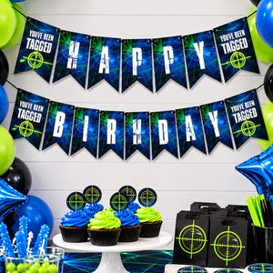 Printable laser tag bunting hangs across a white party backdrop surrounded by blue, lime green and black balloons. Two rows of 5x7" Happy Birthday pennants feature electric laser graphics and bold white gamer lettering, with You've Been Tagged target signs at each end. Coordinating cupcakes, favors and party treats are displayed below. The bright design creates a fun gamer and glow party setting.