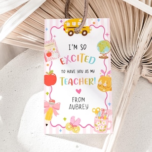 May include: A rectangular gift tag with a white background and pink stripes, featuring a yellow school bus, a globe, a red apple, a pencil with a pink bow, and a floral mug. The text reads: I'M SO EXCITED TO HAVE YOU AS MY TEACHER! FROM AUBREY.