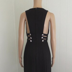 May include: A black evening gown with a high neckline and open back design. The dress features decorative rhinestone buttons and cut-out detailing. The gown is floor-length and displayed on a mannequin.