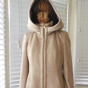 May include: A light beige hooded coat with a full-length zipper. The coat has a tailored fit and a hood that frames the face. The coat is displayed on a mannequin. The hemline of the coat is visible, with a patterned fabric.