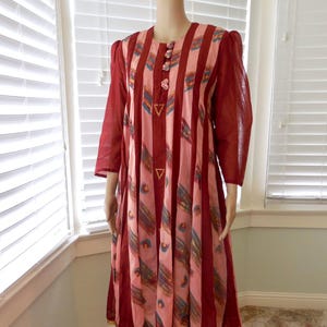 May include: A vintage midi dress with a vertical striped pattern in shades of red and pink. The dress features three-quarter sleeves, decorative buttons, and a flowing skirt. The design includes geometric accents and gold trim.