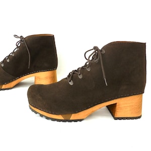May include: A pair of brown suede ankle boots with wooden block heels. The boots have a lace-up design and a rounded toe. The heels are approximately 3 inches high. The soles have a rubber tread for traction.