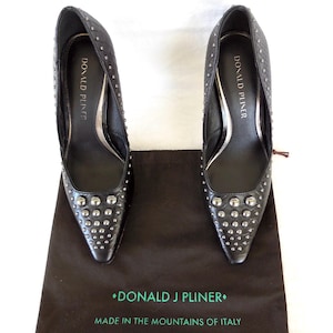 May include: A pair of black pointed-toe pumps with a textured finish and silver-tone metal studs on the toe and sides. The interior insoles are branded with Donald Pliner. A dark brown dust bag features the text Donald J Pliner Made in the Mountains of Italy.