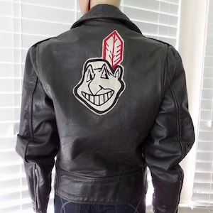 May include: Black leather jacket with a white and red embroidered patch of a cartoon face with a feather on top. The patch is on the back of the jacket.
