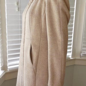 May include: A light beige wool coat with a hood and a large pocket. The coat has a tailored fit and a button closure. The hood is lined with a ribbed knit fabric. The coat is knee-length and suitable for cold weather.