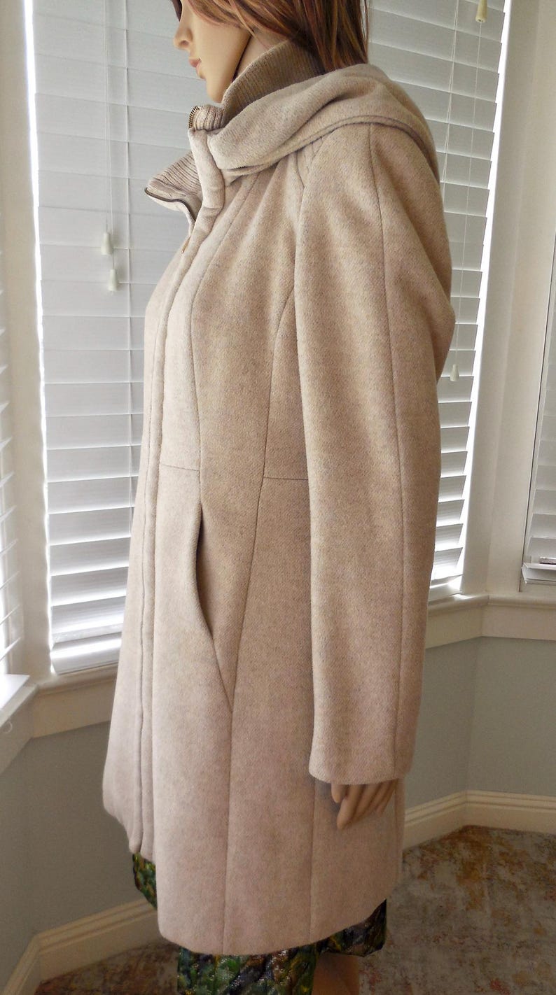 May include: A light beige wool coat with a hood and a large pocket. The coat has a tailored fit and a button closure. The hood is lined with a ribbed knit fabric. The coat is knee-length and suitable for cold weather.