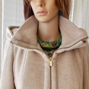 May include: A beige hooded coat with a high collar and a gold zipper. The coat has a knitted collar and a green and gold patterned scarf is visible underneath. The coat is made of a soft, textured material.