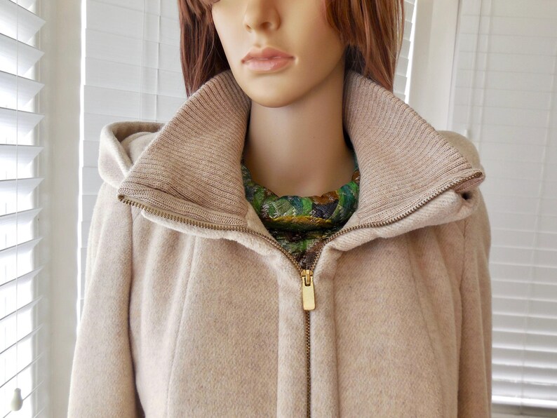 May include: A beige hooded coat with a high collar and a gold zipper. The coat has a knitted collar and a green and gold patterned scarf is visible underneath. The coat is made of a soft, textured material.