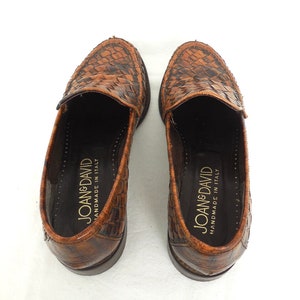 May include: A pair of brown leather loafers with a woven pattern. The loafers are handmade in Italy and have the brand name "Joan & David" printed on the inside.