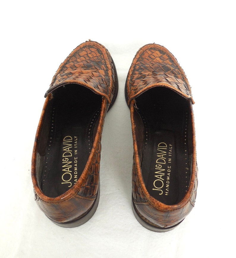 May include: A pair of brown leather loafers with a woven pattern. The loafers are handmade in Italy and have the brand name "Joan & David" printed on the inside.