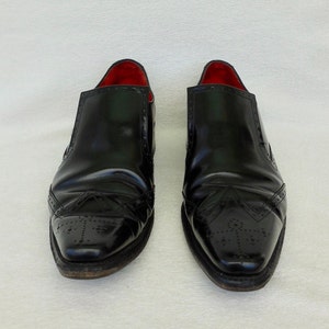 Puede incluir: Un par de zapatos negros de cuero sin cordones con dise&ntilde;o de punta de ala y forro de cuero rojo.