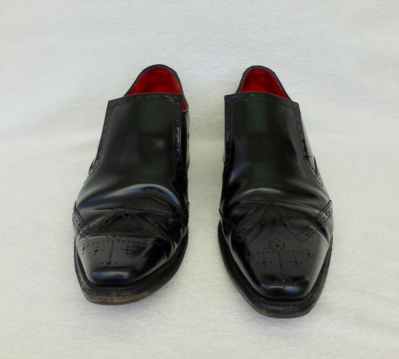 Puede incluir: Un par de zapatos negros de cuero sin cordones con dise&ntilde;o de punta de ala y forro de cuero rojo.