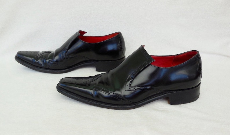 Puede incluir: Un par de zapatos negros de cuero con cordones, punta puntiaguda y detalles de brogue. Los zapatos tienen un forro de cuero rojo.