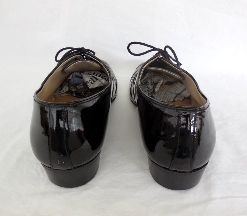 Puede incluir: Par de zapatos de vestir negros de charol con cordones. Los zapatos tienen un acabado brillante y est&aacute;n vac&iacute;os, con papel en el interior. Los zapatos est&aacute;n sobre una superficie blanca.