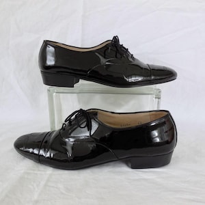Puede incluir: Un par de zapatos Oxford de charol negro con un acabado brillante. Los zapatos tienen un dise&ntilde;o cl&aacute;sico con cierre de cordones y tac&oacute;n bajo. Los zapatos se exhiben sobre un bloque de vidrio transparente sobre un fondo blanco.