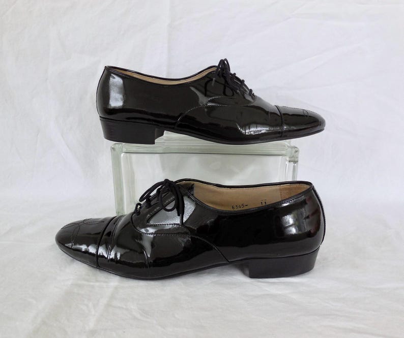 Puede incluir: Un par de zapatos Oxford de charol negro con un acabado brillante. Los zapatos tienen un dise&ntilde;o cl&aacute;sico con cierre de cordones y tac&oacute;n bajo. Los zapatos se exhiben sobre un bloque de vidrio transparente sobre un fondo blanco.