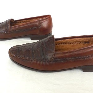 K&ouml;nnte beinhalten: Ein Paar braune Lederschuhe mit Kroko-Pr&auml;gung. Die Loafer haben eine gen&auml;hte Kante und eine spitze Zehenpartie.