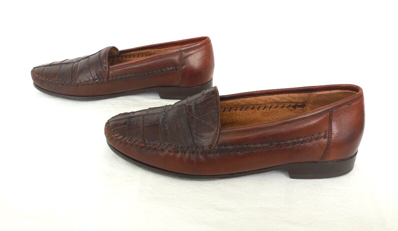 K&ouml;nnte beinhalten: Ein Paar braune Lederschuhe mit Kroko-Pr&auml;gung. Die Loafer haben eine gen&auml;hte Kante und eine spitze Zehenpartie.