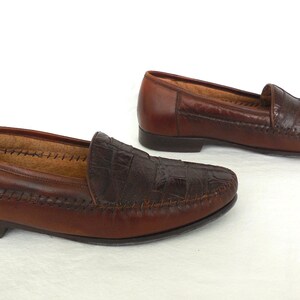 K&ouml;nnte beinhalten: Ein Paar braune Lederschuhe mit Kroko-Pr&auml;gung. Die Loafer haben eine gestickte Kante und eine spitze Zehenpartie.