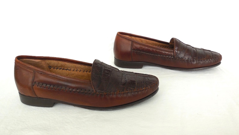 K&ouml;nnte beinhalten: Ein Paar braune Lederschuhe mit Kroko-Pr&auml;gung. Die Loafer haben eine gestickte Kante und eine spitze Zehenpartie.