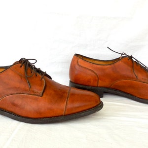 May include: A pair of brown leather dress shoes with dark brown laces. The shoes have a classic cap-toe design and a dark sole. The shoes are displayed on a white surface.