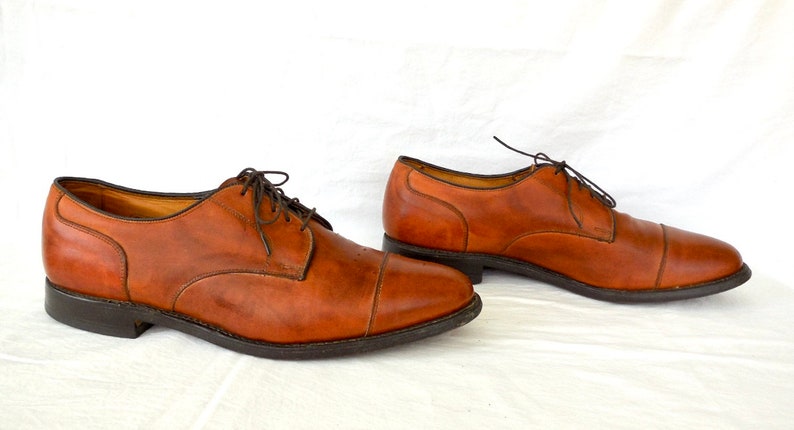 May include: A pair of brown leather dress shoes with dark brown laces. The shoes have a classic cap-toe design and a dark sole. The shoes are displayed on a white surface.