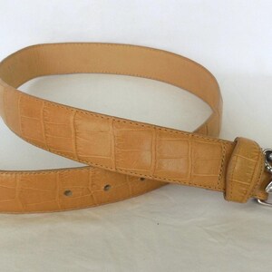 May include: Tan leather belt with a silver decorative buckle. The belt has a textured, crocodile-like pattern. The buckle features a detailed leaf design. A stylish accessory for any outfit.