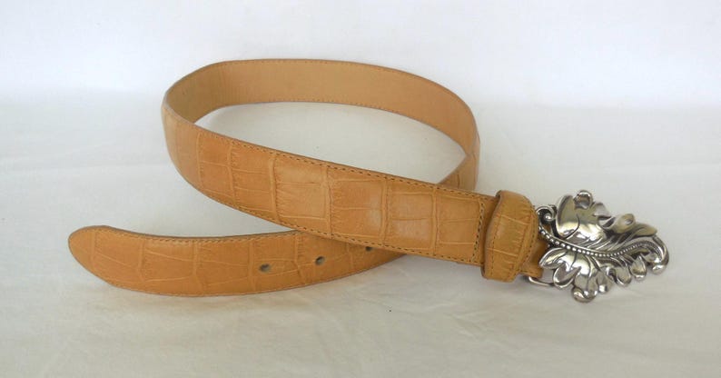 May include: Tan leather belt with a silver decorative buckle. The belt has a textured, crocodile-like pattern. The buckle features a detailed leaf design. A stylish accessory for any outfit.