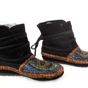 K&ouml;nnte beinhalten: Paar schwarze Wildleder-Ankle Boots mit Perlenbesatz. Die Stiefel haben einen Wickelverschluss und sind mit aufwendigen Perlenarbeiten in Blau, Gr&uuml;n und Kupfer verziert. Die Sohlen sind schwarz.