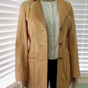 May include: A tan leather jacket with decorative stitching and button closures. The jacket has a long, fitted silhouette.